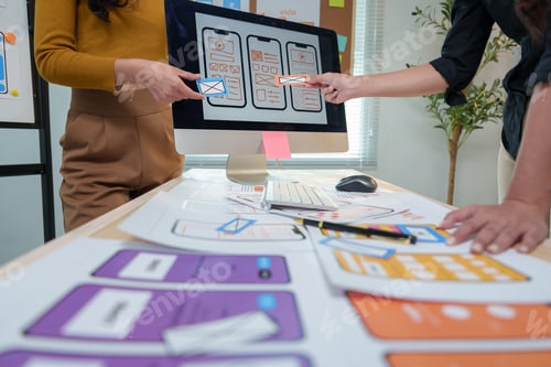 Preview: Two ux designers are comparing mobile application wireframe sketches on a desktop computer, analyzin