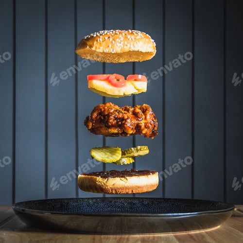 Preview: Vertical shot of a spicy chicken burger with gherkins, pineapple, and chili falling in stacks