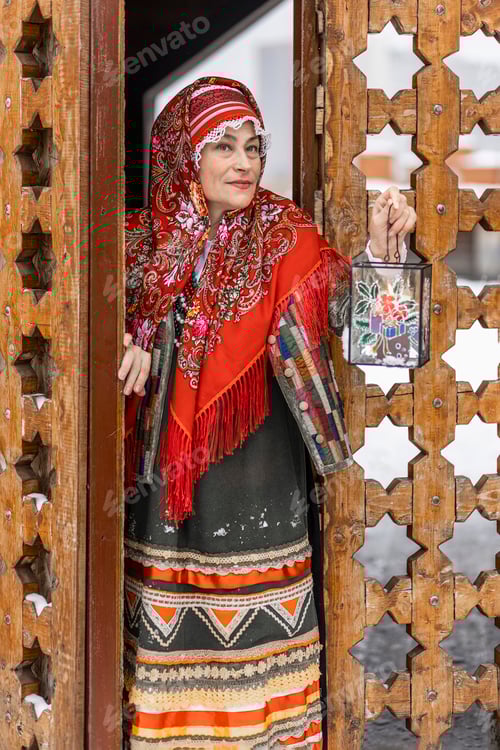 Preview: A elderly woman in a national Slavic costume looks out of wooden vintage doors, shining a lantern
