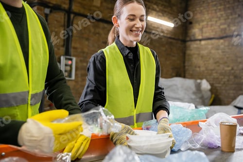 Preview: Positive young worker in gloves and protective vest holding plastic trash near blurred colleague
