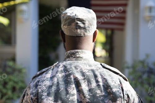 Preview: Rear view of african american male soldier standing in front of american flag outside home