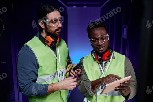 Preview: Two network technicians using tablet while repairing server in neon light