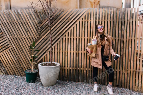 Preview: Full-length portrait of stylish young woman with camera posing in front of wooden fence after shoppi