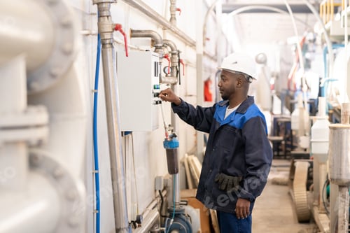 Preview: Young African male in workwear standing by industrial machine and turning knob on control panel