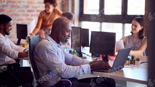 Preview: Mature Businessman Working On Laptop At Desk In Busy Multi-Cultural Office