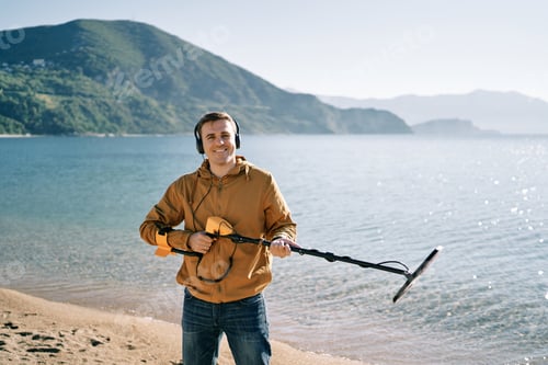 Preview: Smiling man with a metal detector at the ready stands on the seashore