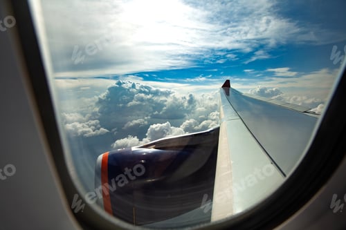 Preview: Scenic view of airplane wing and engine through passenger window flying above clouds under blue sky
