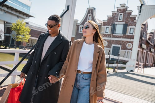Preview: happy and fashionable couple in sunglasses holding hands while walking with shopping bags across