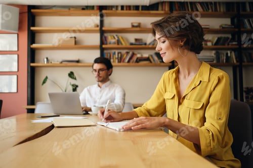 Preview: Young woman sitting at the table happily writing in notepad with colleague on background in office
