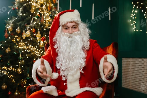 Preview: Santa Claus with outstretched arms looking at the camera, by the Christmas tree.