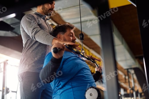 Preview: Mature man working out on exercise machine with coach's assistance in a gym.