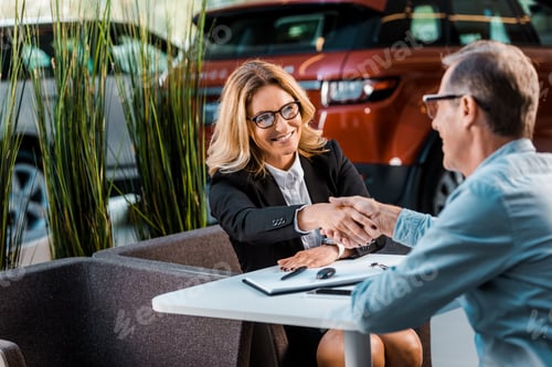 Preview: happy adult customer and female car dealer shaking hands in showroom