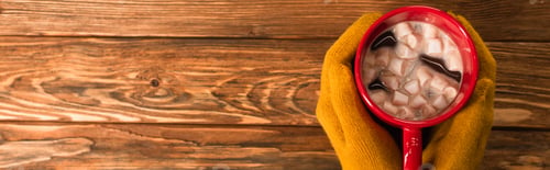 Preview: cropped view of person in yellow gloves holding cup of cocoa with marshmallows above wooden surface,
