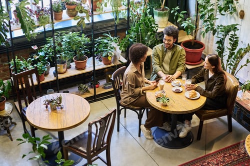 Preview: Happy young man and two intercultural women chatting by table in cafe