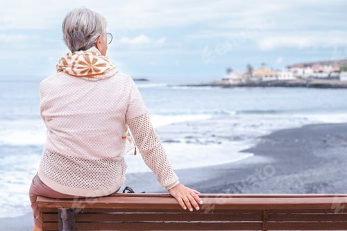 Preview: Rear view of senior woman wearing eyeglasses sitting on outdoors bench at sea looking away