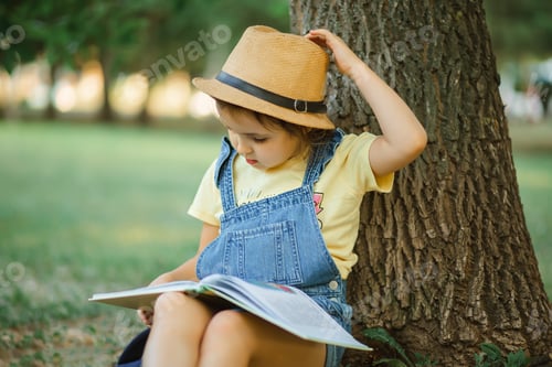 Preview: Little cute girl kid sitting under the tree on summer in the park outside and reading a book.