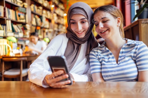Preview: Two teenage girls of different nationalities are sitting in library and looking at phone