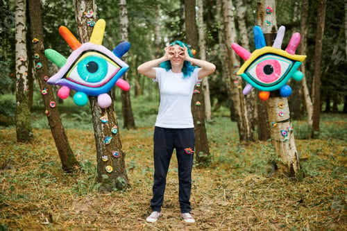 Preview: Weird young adult woman with dyed turquoise hair in white T shirt near big eyes art installation