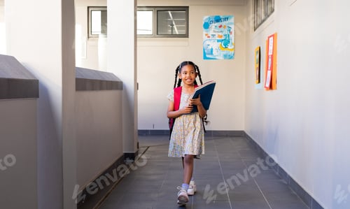 Preview: Happy biracial schoolgirl with books and school bag at elementary school corridor
