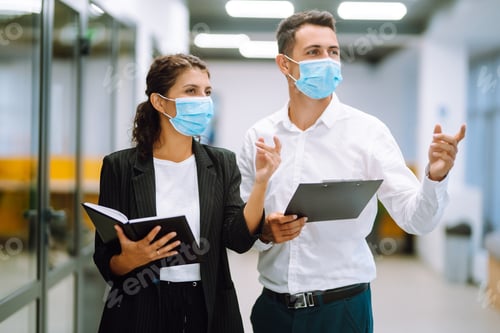 Preview: Two colleagues in protective masks in office discussing together work issues standing near elevator.