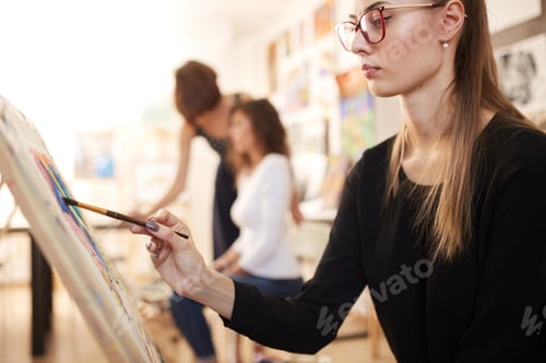 Preview: Nice young girl in glasses dressed in black blouse and jeans sits at the easel and paints a picture