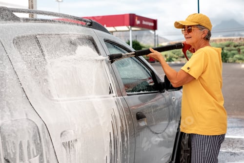 Preview: Smiling caucasian senior woman washing her car in a self-service car wash station using high pressur