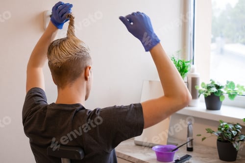 Preview: Young caucasian male with blue gloves applying bleaching dye on hairs in front of mirror at home