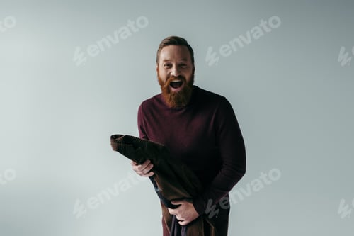 Preview: excited and bearded man in turtleneck holding blazer while looking at camera isolated on grey