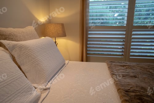 Preview: white pillows on bed with side lamp on, with natural light coming in from the window