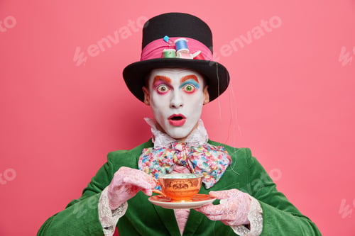 Preview: Horizontal shot of stupefied crazy man has image of hatter drinks hot tea poses with cup wears hat a