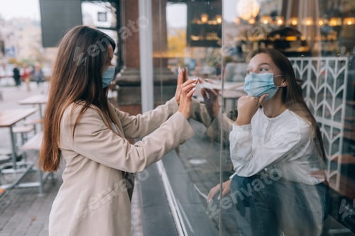Preview: Two women in protective masks opposite each other, window between them