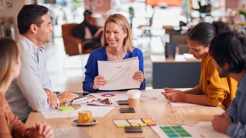 Preview: Businesswoman Giving Presentation To Colleagues Sitting Around Table In Modern Open Plan Office