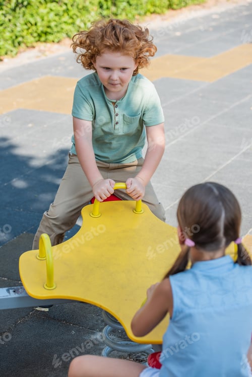 Preview: curly boy riding seesaw with blurred sister outdoors