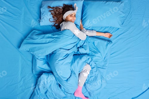 Preview: Beautiful brunette young woman with happy expression lying on comfy bed alone under soft blanket dre