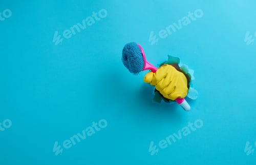 Preview: Hand in a yellow rubber glove holds a plastic cleaning brush on a blue background