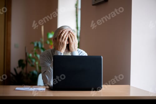 Preview: a lonely old man with a gray beard hides his face in his palms sitting at a laptop computer at home