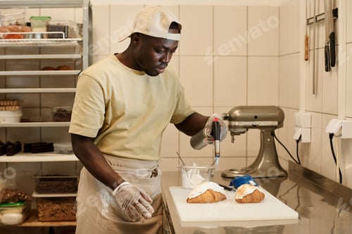 Preview: Young Black man as male baker decorating fresh croissants with cream in bakery