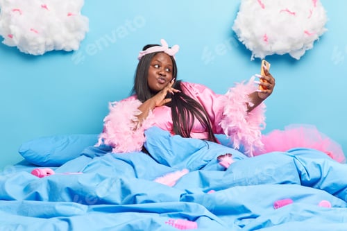 Preview: Attractive dark skinned chubby lady pouts lips at smartphone camera makes selfie portrait wears pink