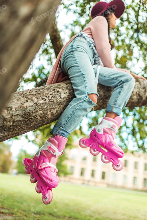 Preview: low angle view of stylish young woman in roller skates sitting on tree in park