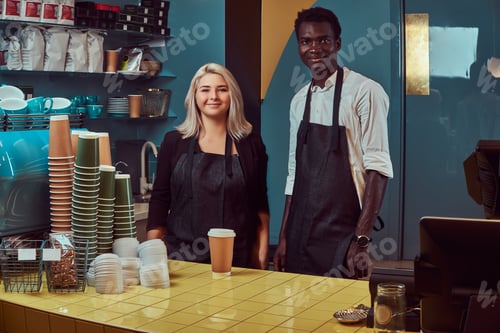 Preview: Two young multiracial baristas in aprons standing welcomingly at their trendy coffee shop.