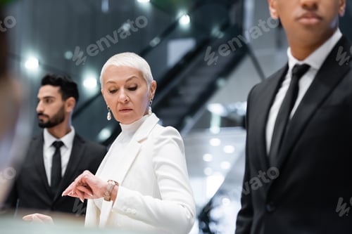 Preview: mature businesswoman checking time on wristwatch near interracial bodyguards in hotel