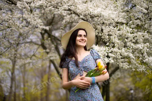 Preview: A young girl in a hat and with tulips, standing in a blue dress next to a blooming white tree