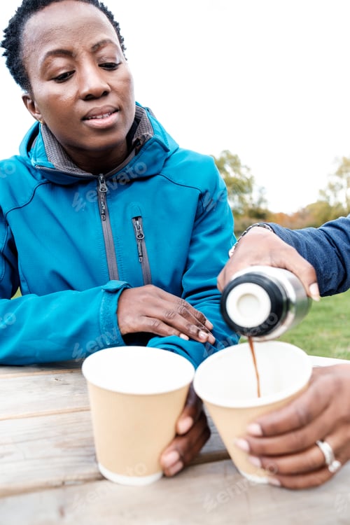 Preview: Unrecognizable black woman serving coffee to her female friend in a park.