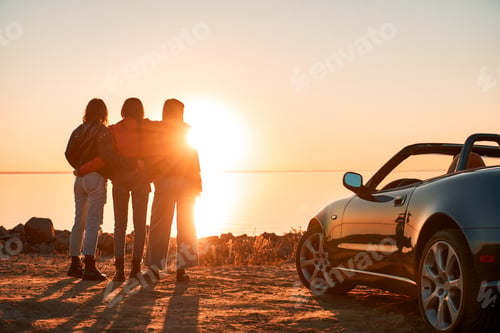 Preview: Cheerful young three women standing near cabriolet wathing at sunset