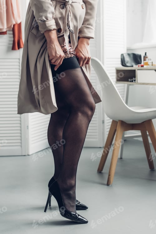 Preview: cropped shot of transgender woman in trench coat wearing stockings at home