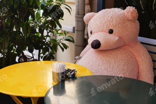 Preview: Large pink bear sits at a round table outside in the sun