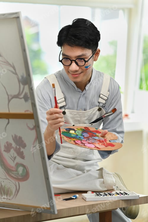Preview: Happy asian male artist holding paintbrushes and palette painting on canvas at art studio.
