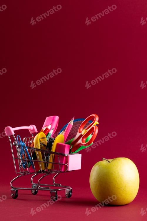 Visualização: Vista de perto do carrinho de compras em miniatura com materiais escolares, maçã e espaço de cópia em fundo vermelho