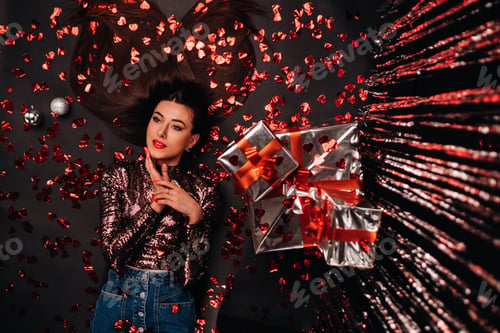 Preview: Top view of a girl lying in shiny clothes on the floor in confetti in the form of hearts and gifts