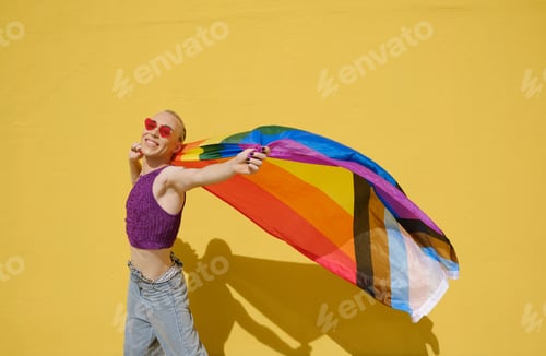 Preview: Young stylish non-binary person carrying the lgbt rainbow flag against a yellow wall.